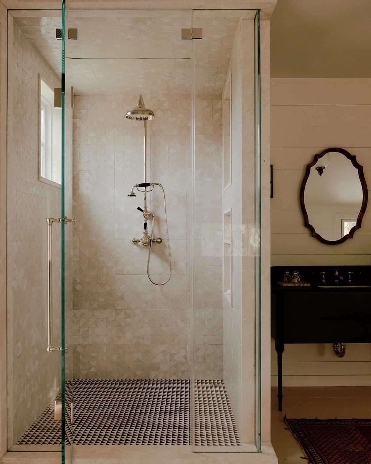 Glass-enclosed shower with cream diamond-pattern zellige tiles, chrome fixtures, and black mosaic floor beside dark steel vanity