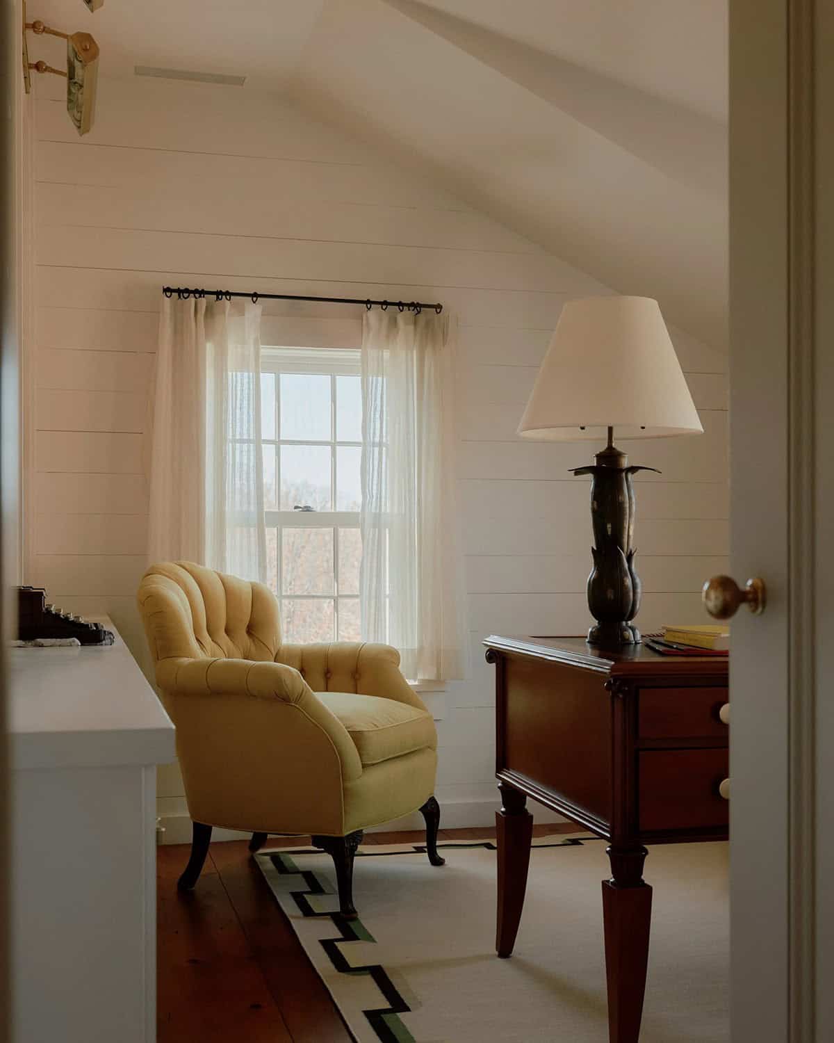 Attic bedroom corner with tufted yellow armchair, antique mahogany dresser, table lamp, and sloped shiplap ceiling