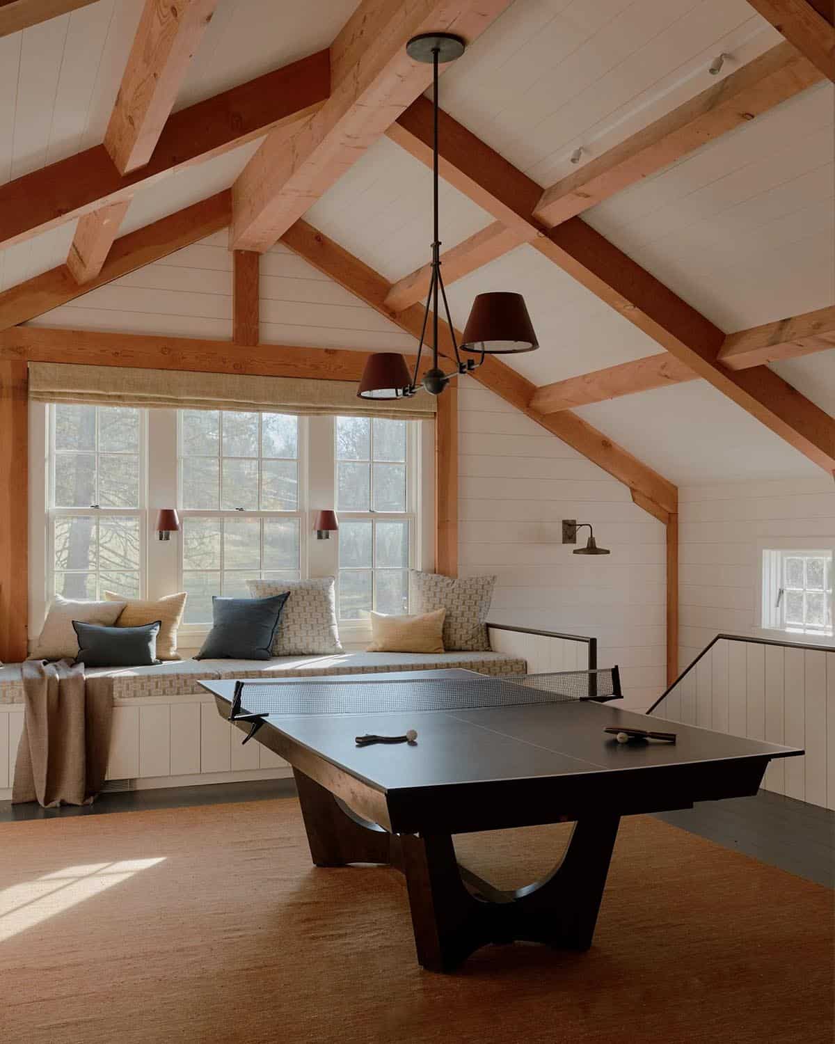 Barn game room with ping pong table, built-in window seat, exposed timber truss ceiling, and burgundy pendant lights