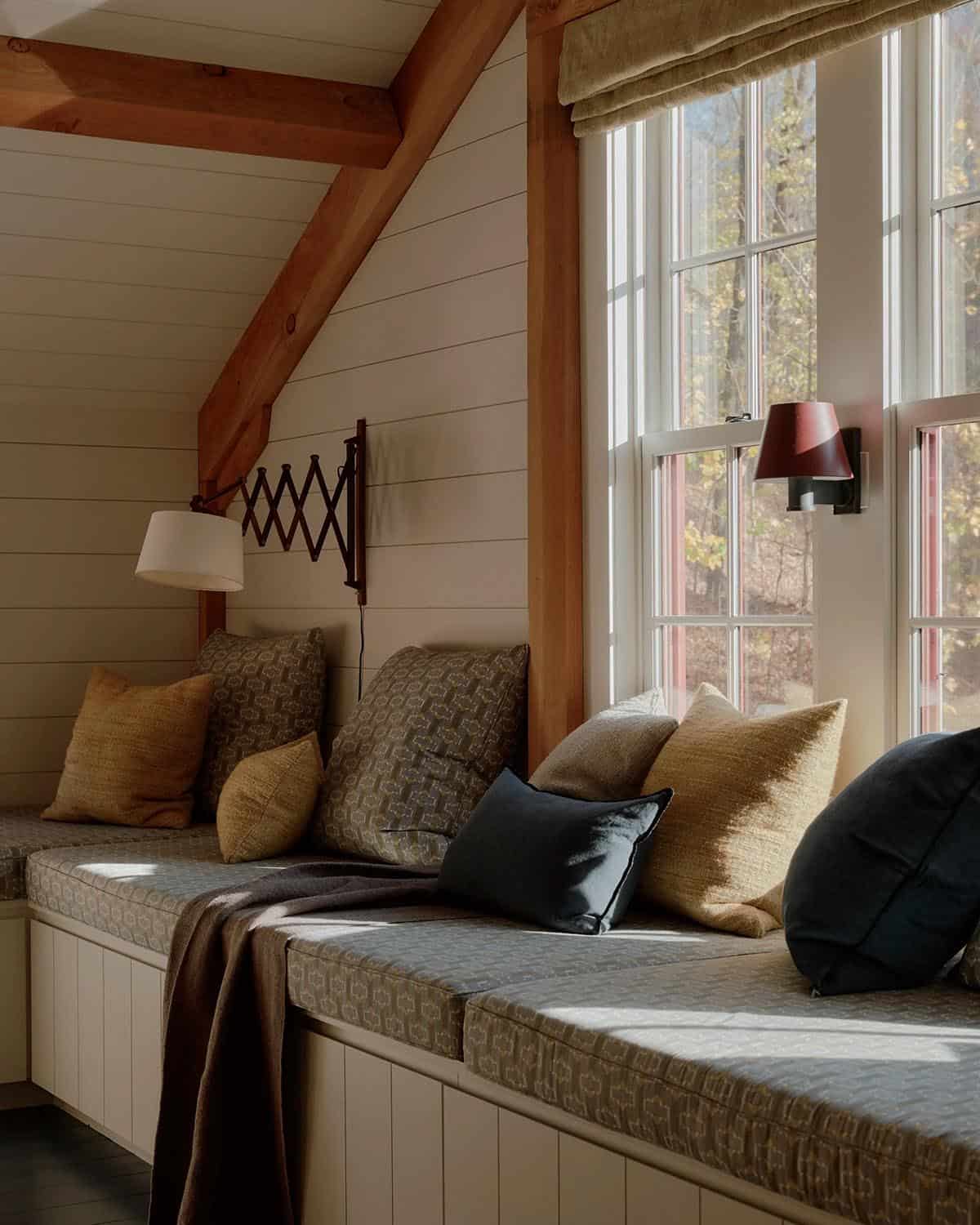 Built-in window seat nook with mixed pattern cushions and pillows, exposed timber beam, accordion wall lamp, and red barn sconces