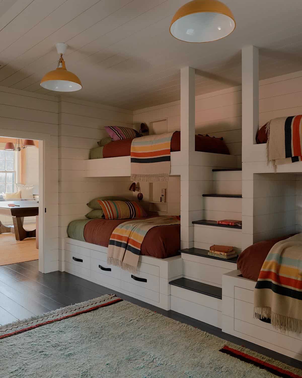 Bunk room with four built-in bunks, colorful striped blankets, under-bed storage drawers, and yellow dome pendants