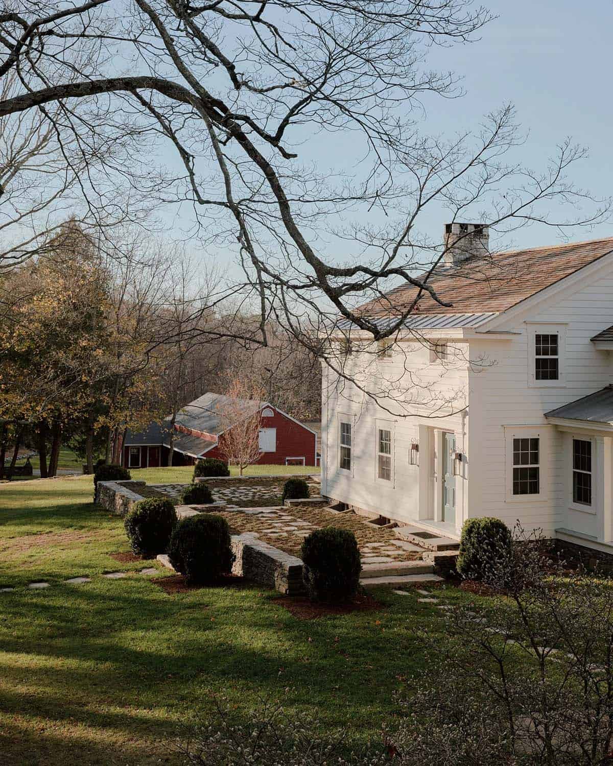 White farmhouse exterior with columned entry porch, boxwood landscaping, stone path, greenhouse, and red barn beyond
