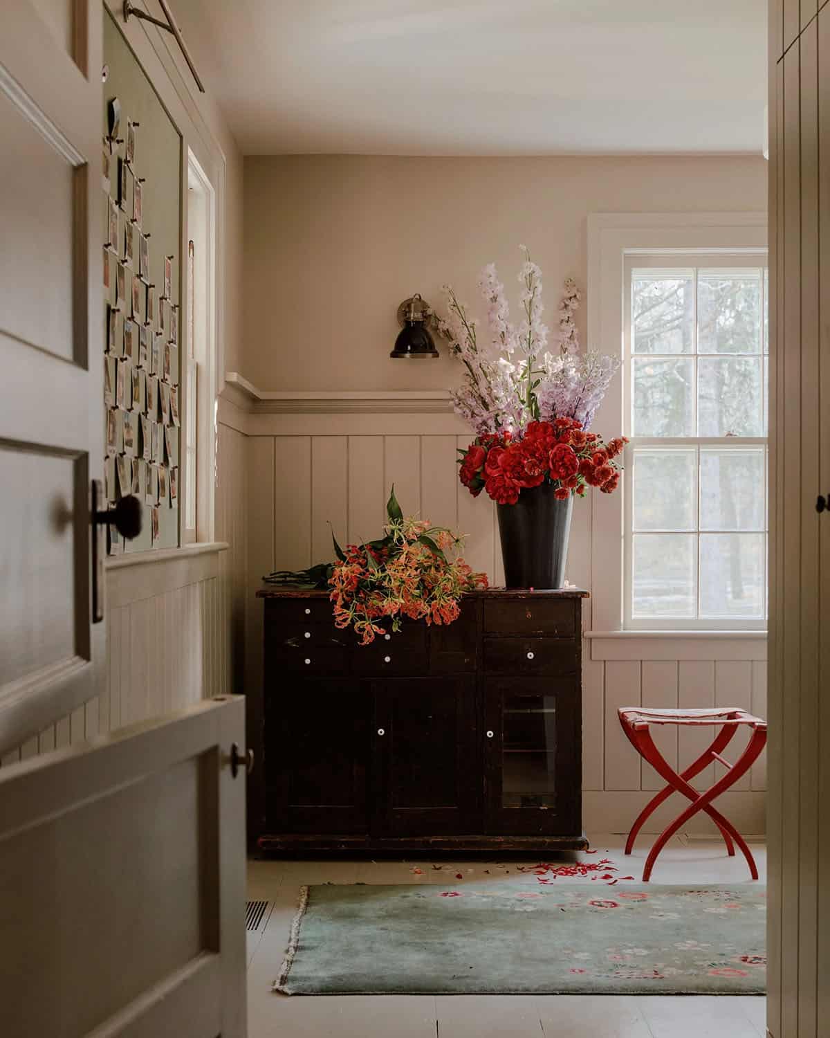 Mudroom or utility room with large floral arrangement on antique cabinet, red folding stool, and photo display board