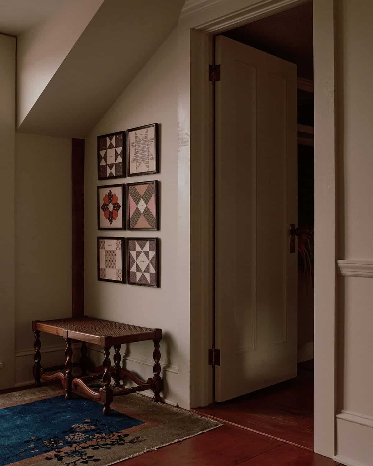 Dark hallway corner with six framed quilt block prints in a grid, antique cane bench, and Persian rug