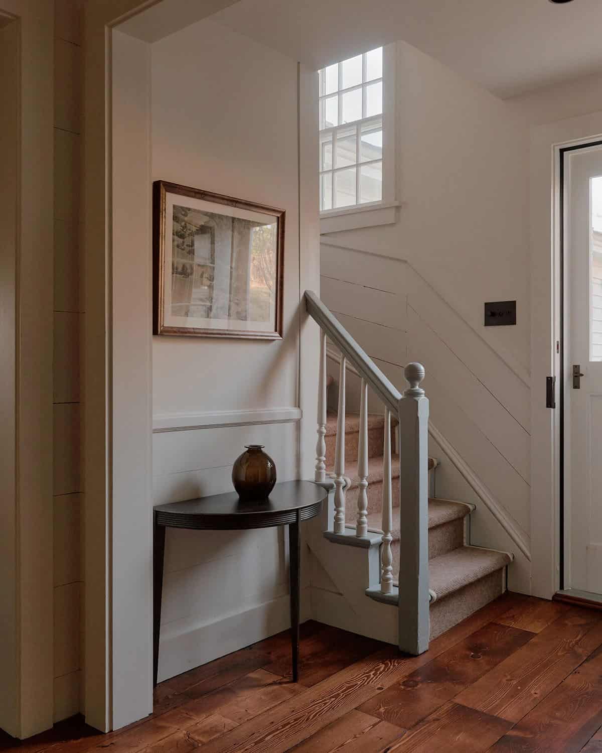 Farmhouse entry with painted staircase, demilune console table, amber vase, framed landscape print, and wide-plank floors