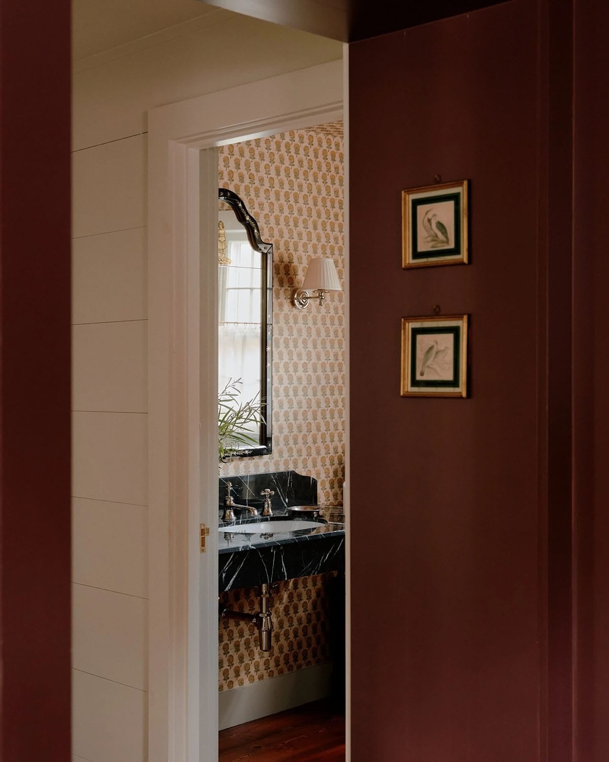 View through burgundy-painted doorway into powder room with block-print wallpaper and black marble vanity, with framed bird prints beside