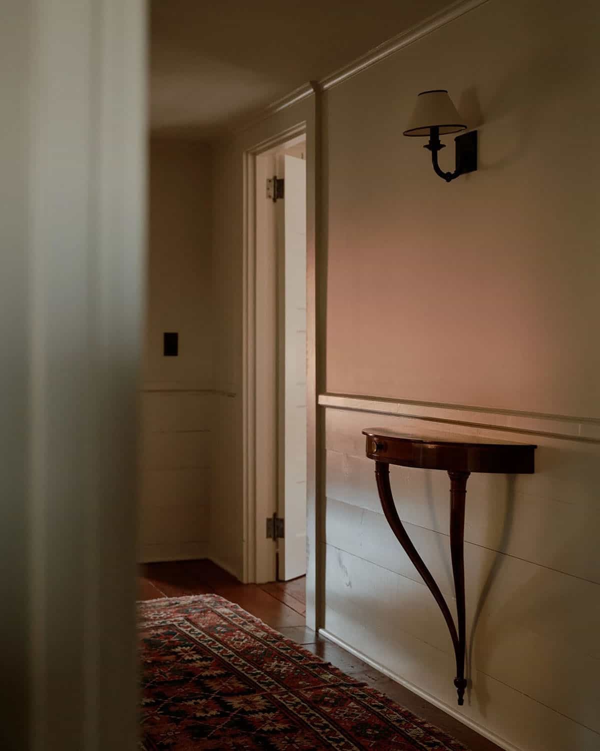 Dim hallway with antique demilune console table, iron sconce, Persian runner, and open door to shiplap room beyond