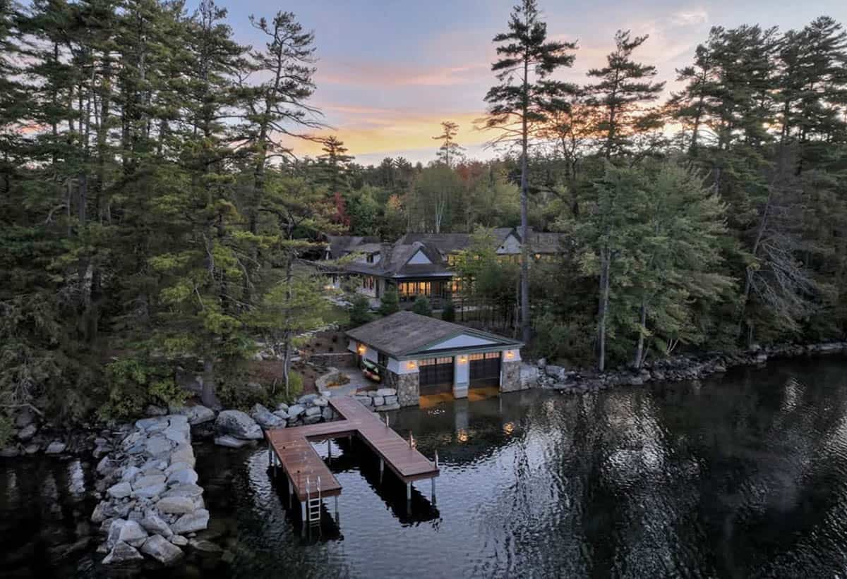Aerial sunset view of lit boathouse, wooden dock, and main house through pine forest
