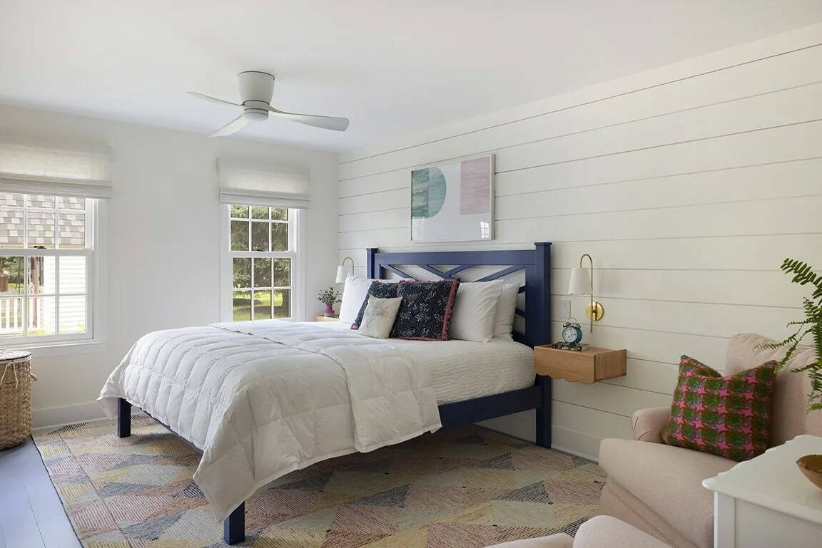 Bright bedroom with navy blue bed frame, shiplap accent wall, gold sconce, and pink armchair with colorful rug