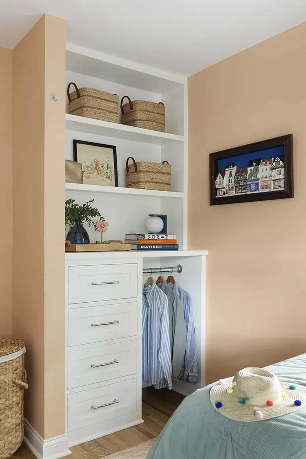 Peach bedroom with white built-in closet, open shelving with wicker baskets, and art books on display