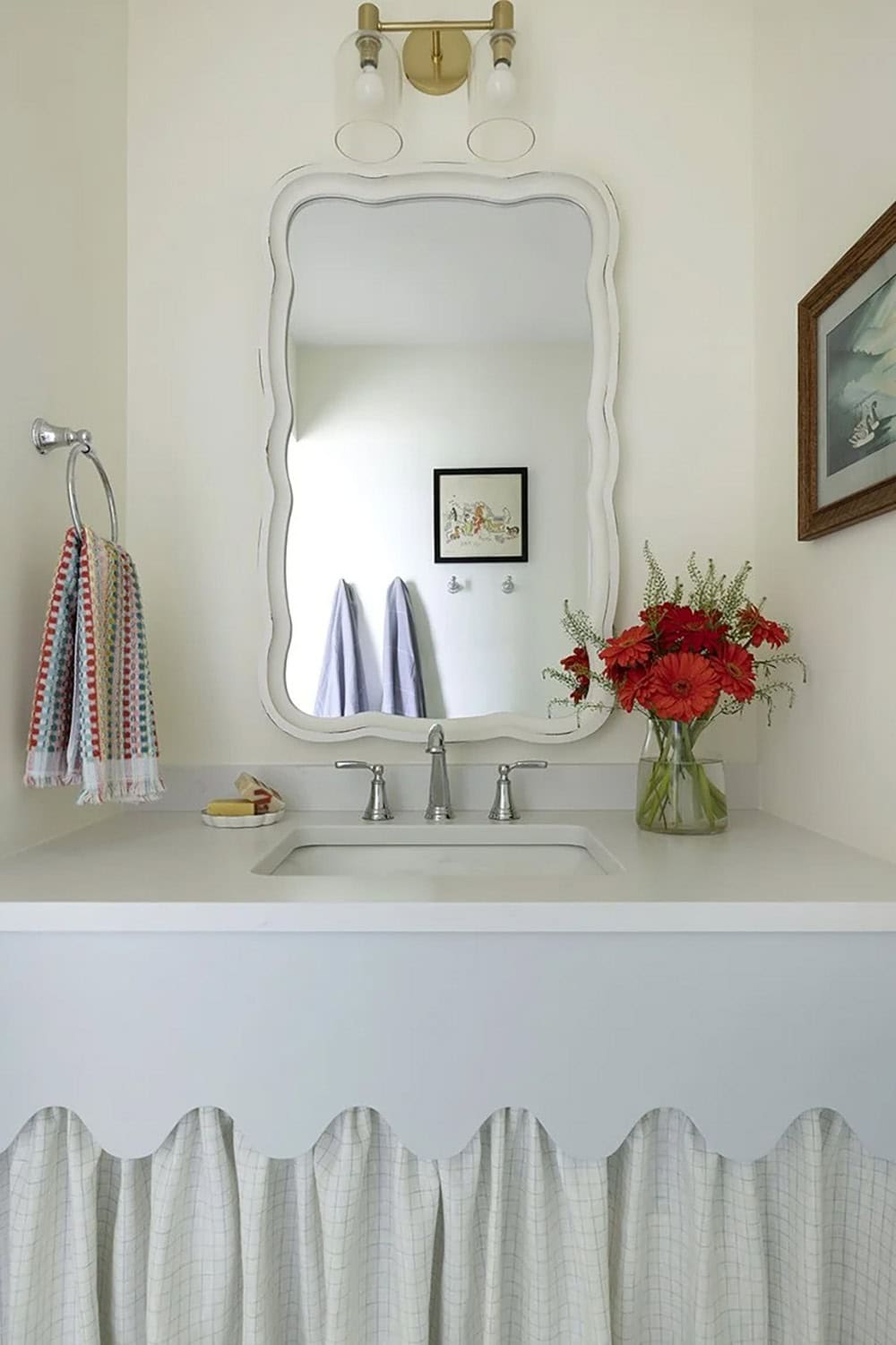 Bathroom vanity with wavy white mirror frame, gold bar sconce, chrome fixtures, and skirted under-sink storage
