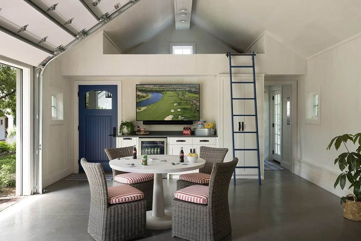 Converted garage game room with wicker chairs, round white table, wall-mounted TV, blue barn door, and library ladder