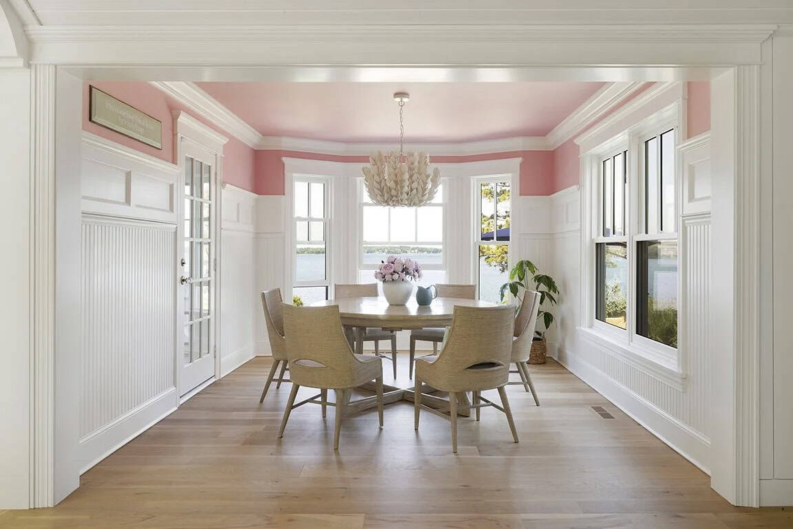 Formal dining room with pink painted ceiling, wainscoting, round wood table, rattan chairs, and leaf chandelier with lake views
