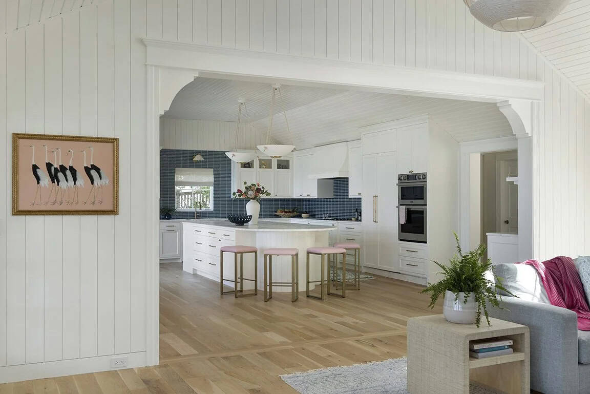 View from shiplap great room through arched opening into white kitchen with curved island, pink stools, and blue tile backsplash