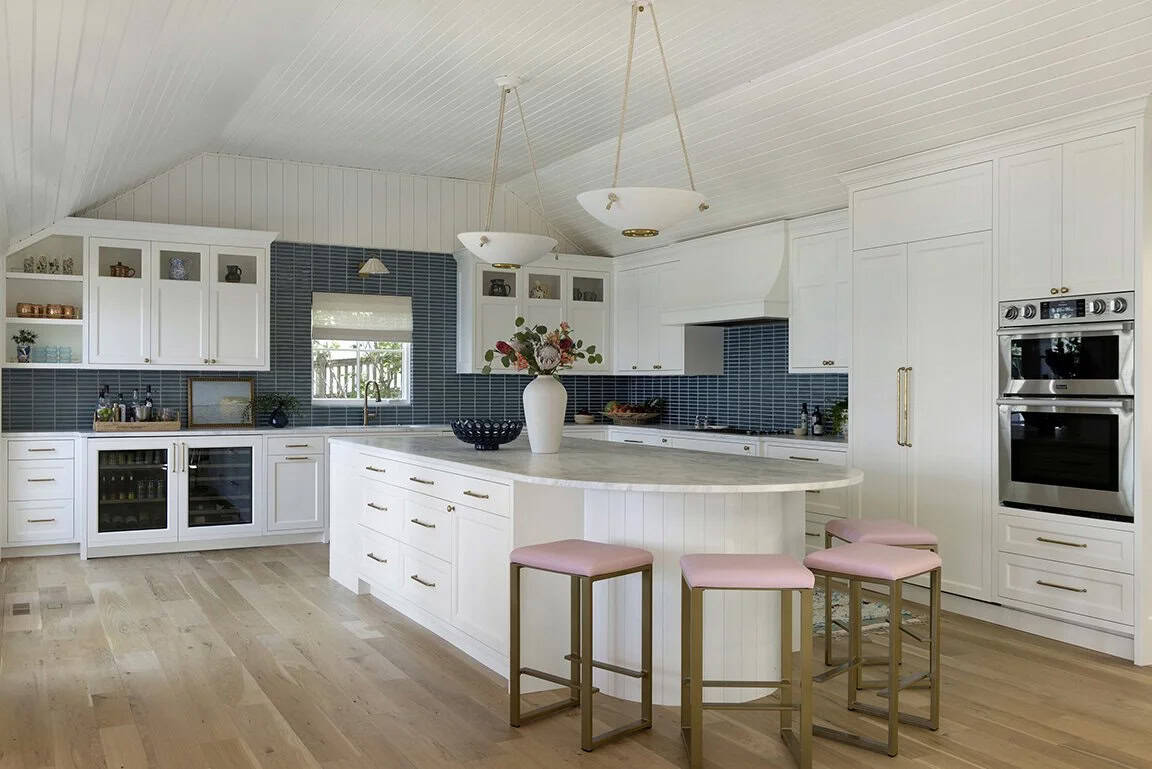 White kitchen with curved oval island, pink upholstered gold stools, blue stacked tile backsplash, and glass-front upper cabinets
