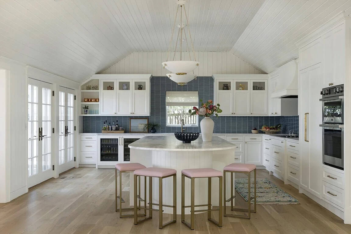 Spacious white kitchen with vaulted shiplap ceiling, oval island, pink bar stools, blue tile backsplash, and French doors