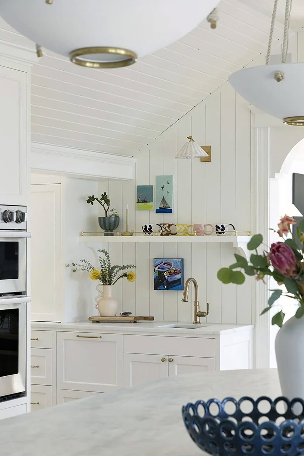 Kitchen detail with brass floating shelves, small oil paintings, gold sconce, white cabinetry, and prep sink on shiplap wall