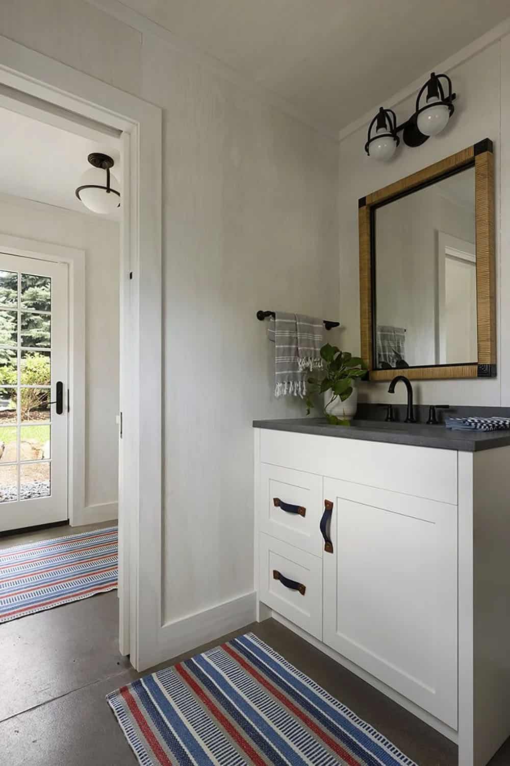 Garage bathroom with white shaker vanity, leather pulls, rattan-framed mirror, black globe sconces, and red-white-blue striped rug