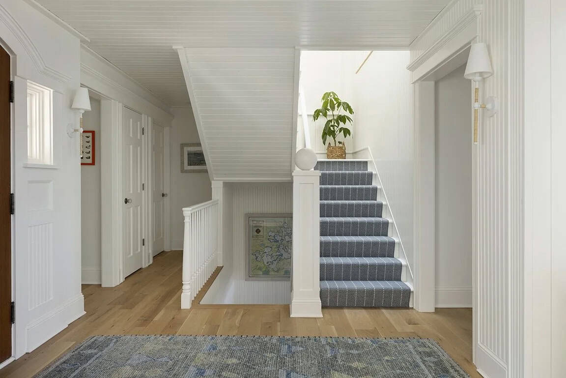 White cottage foyer with beadboard ceiling, blue patterned stair runner, white balusters, framed map art, and wall sconces