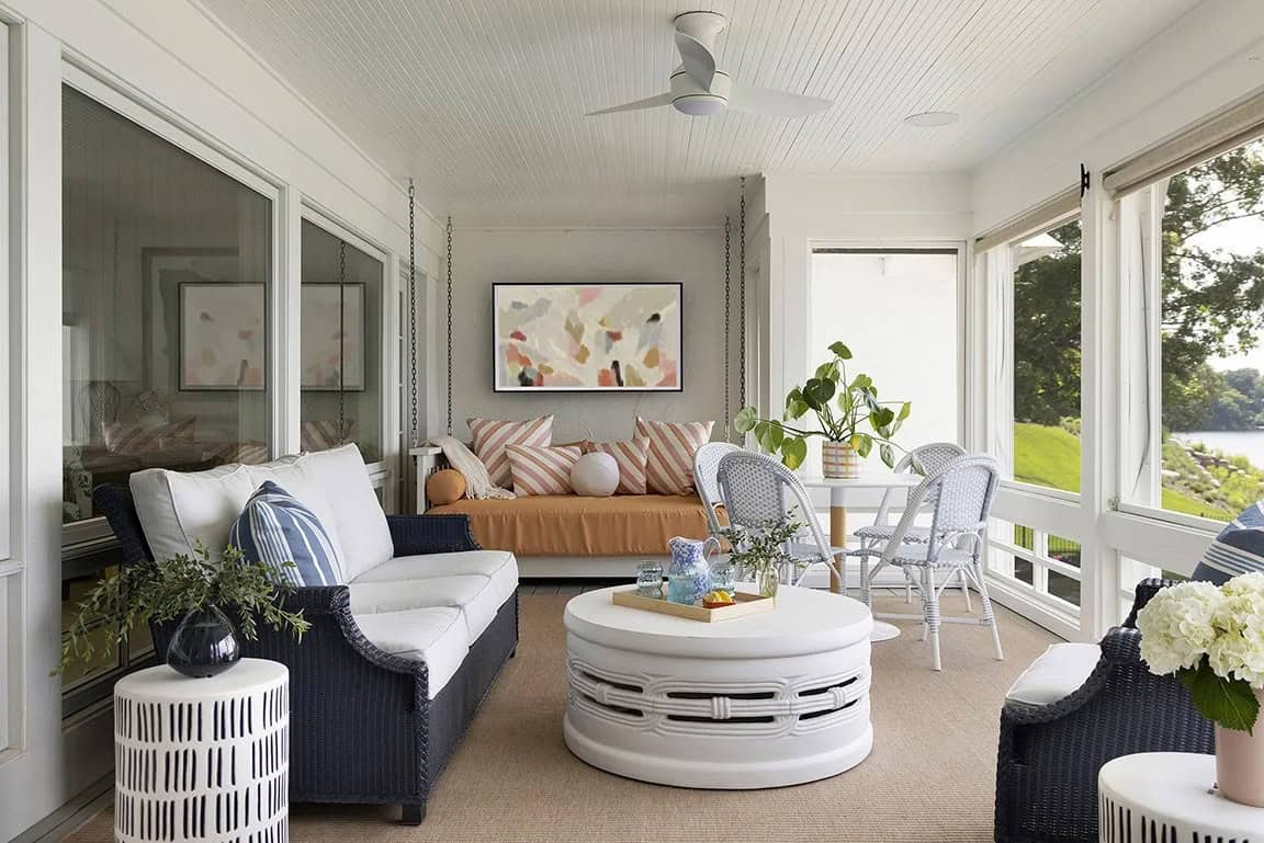 Lakeside screened porch with navy wicker furniture, white drum coffee table, hanging daybed with striped pillows, and abstract art
