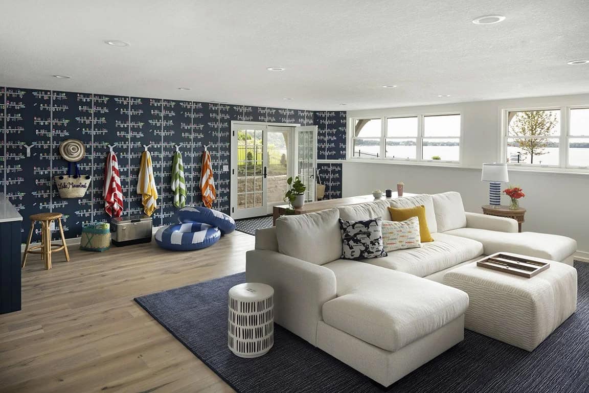 Basement lake room with navy boat wallpaper, white sectional sofa, colorful beach towels on hooks, and lake views through windows