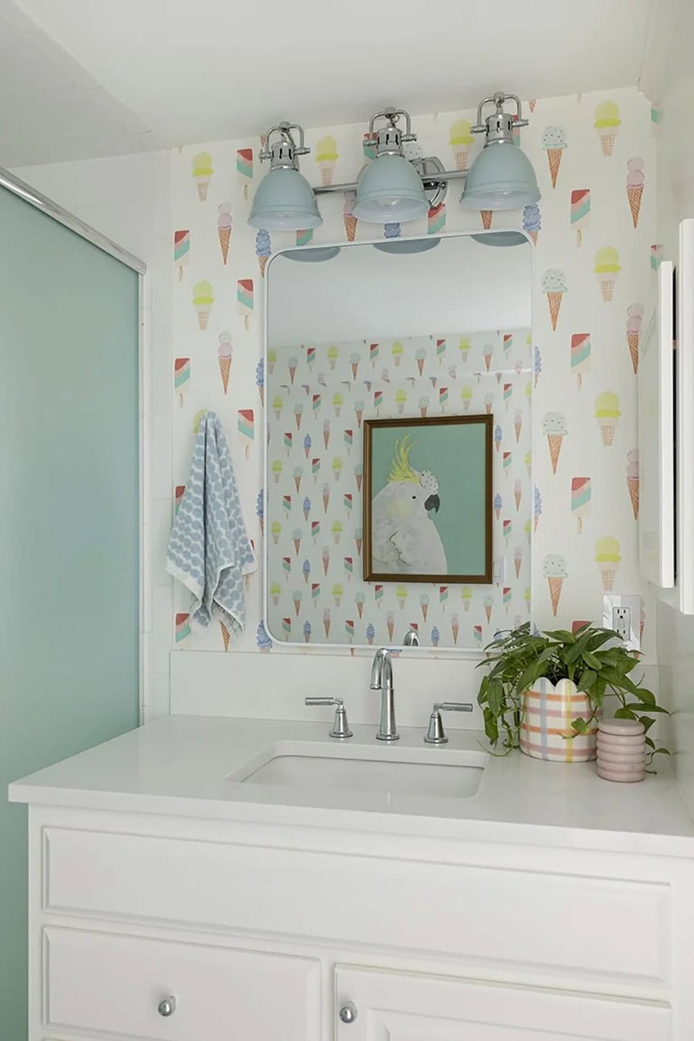 Playful bathroom with ice cream cone wallpaper, blue bell sconces, white vanity, and cockatoo artwork above the sink