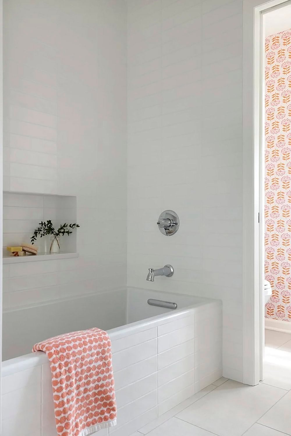 Clean white tiled tub surround with built-in niche, chrome fixtures, coral towel, and floral wallpaper glimpsed through doorway