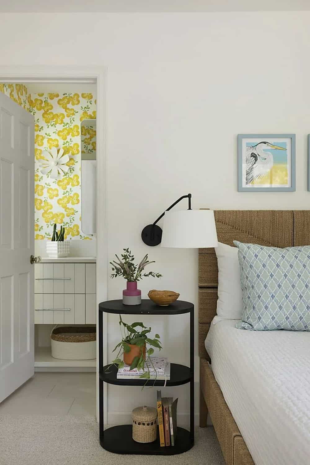 Guest bedroom nightstand vignette with black round tiered table, woven headboard, heron art, and yellow floral en-suite beyond