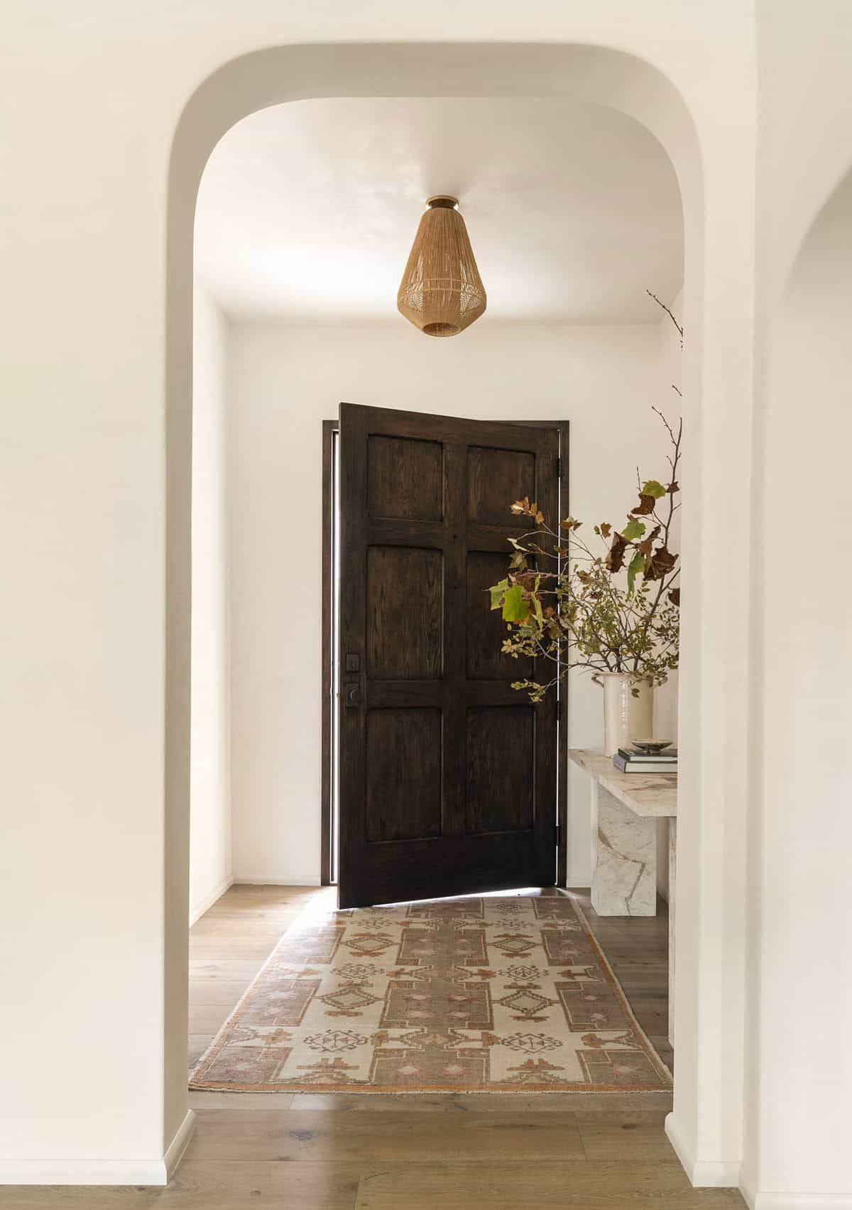 Entryway with rounded plaster arch, dark wood front door, woven pendant light, and vintage runner rug