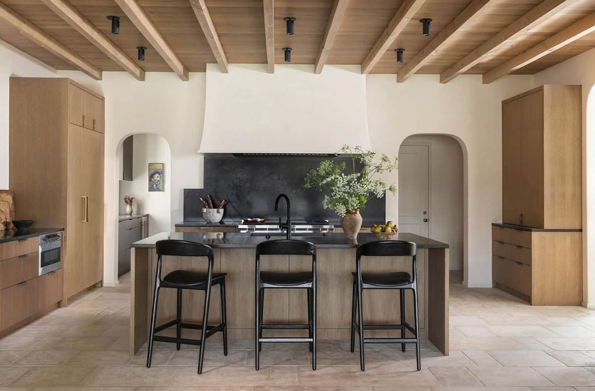 Kitchen with white oak cabinetry, exposed wood beam ceiling, black stone island, and arched pass-throughs