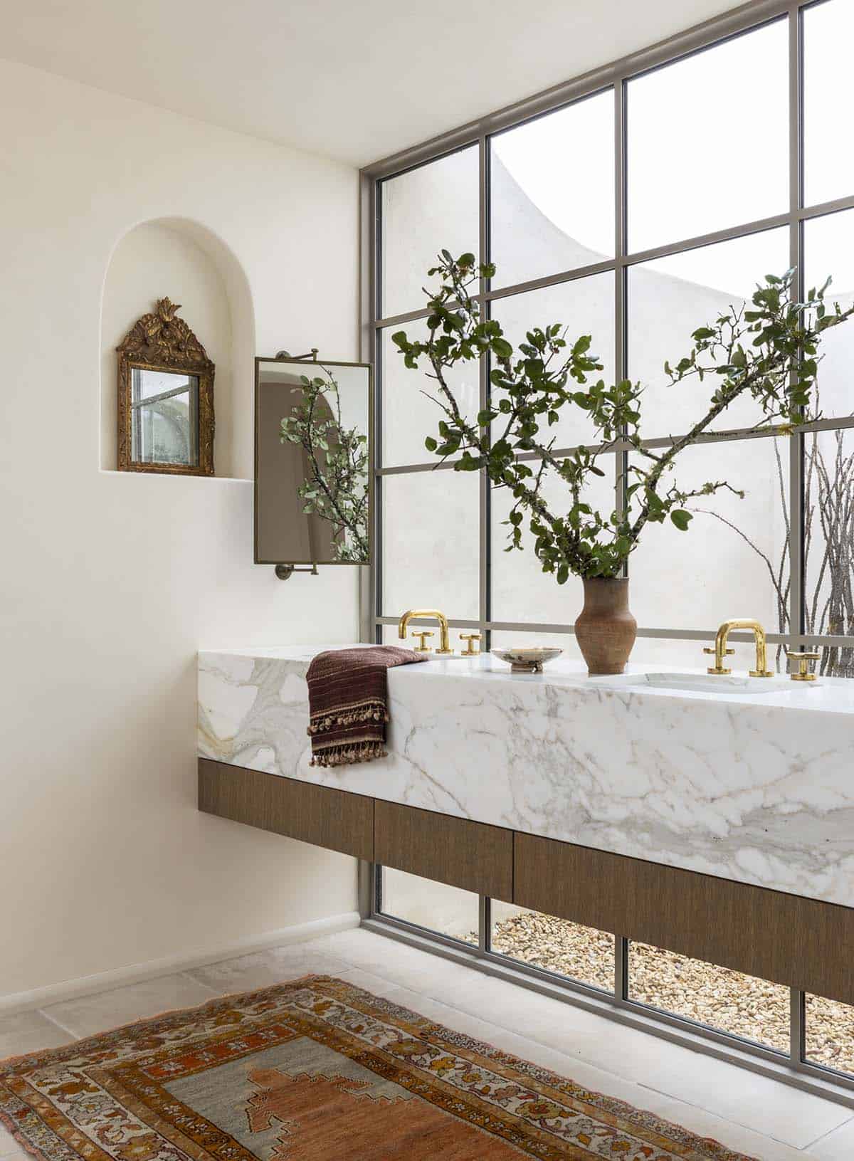 Primary bathroom with floating marble double vanity, brass faucets, arched wall niche, and steel-framed window