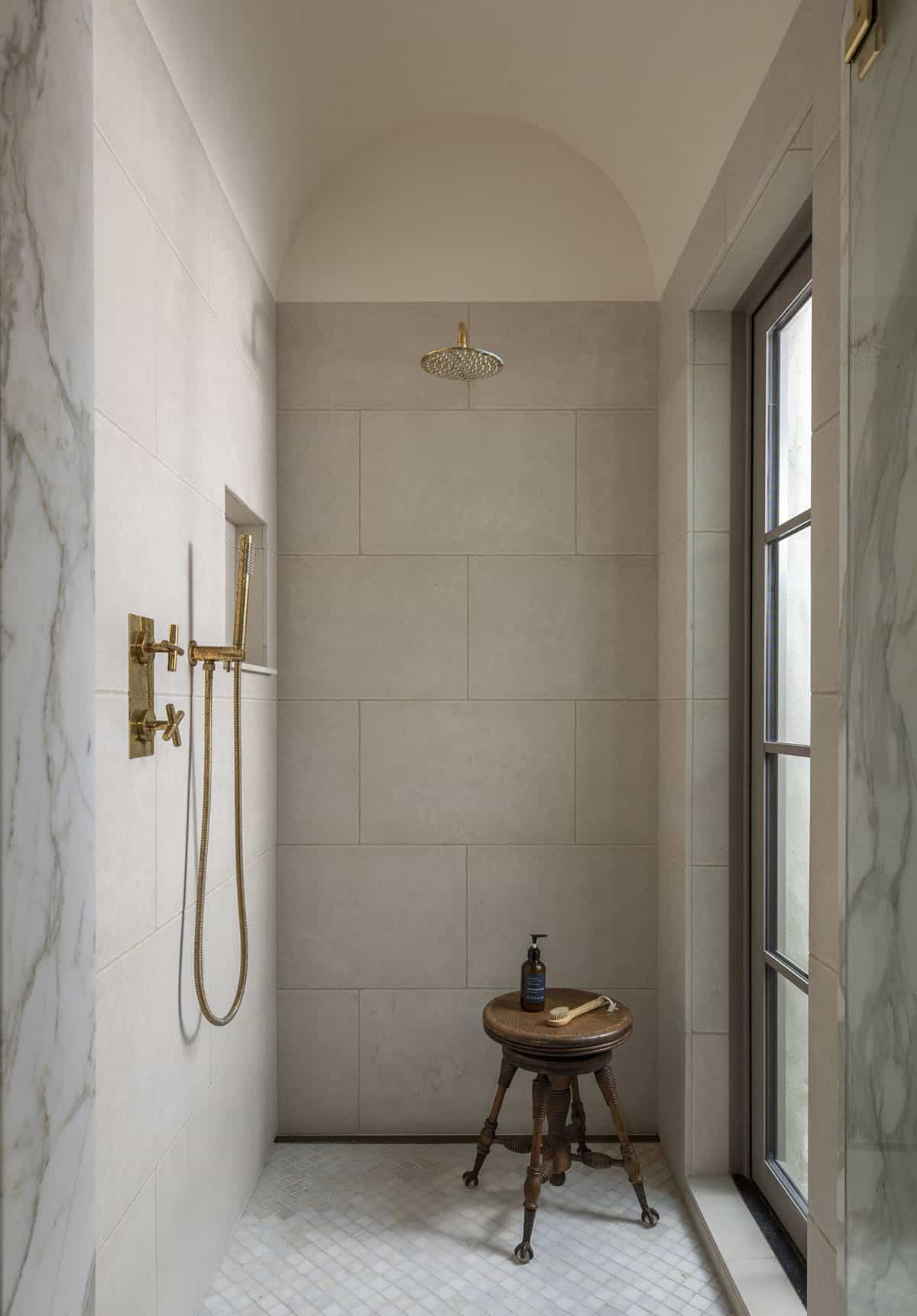 Walk-in shower with arched ceiling niche, brass rainfall head, limestone tile walls, and antique wood stool