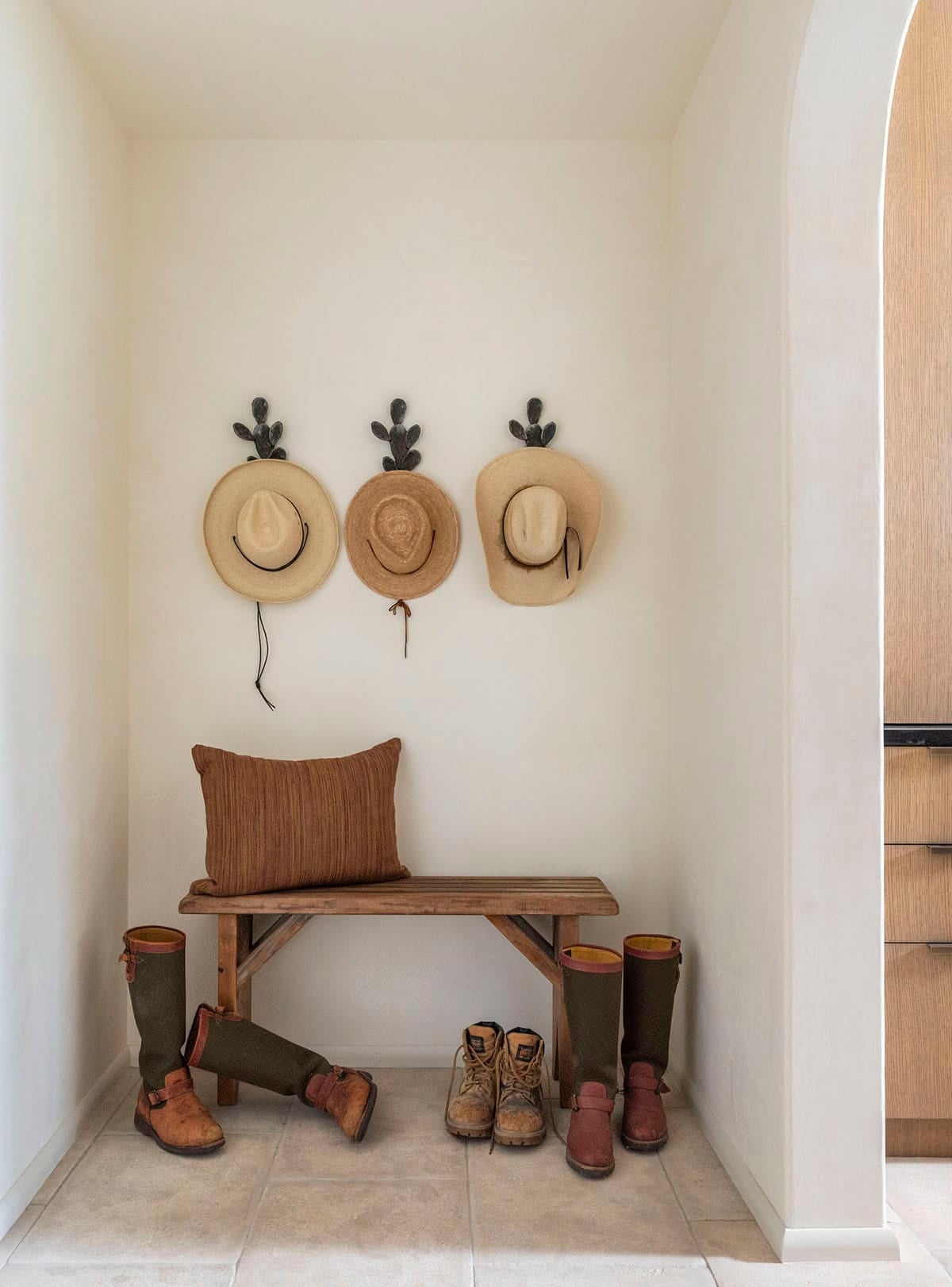 Mudroom niche with three straw hats on cactus-shaped hooks, rustic wood bench, and riding boots below
