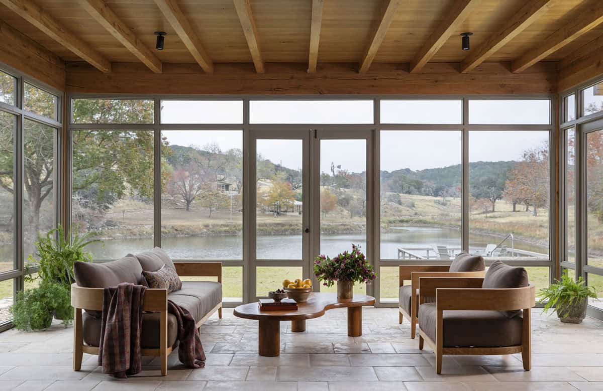 Glass-enclosed sunroom with wood beam ceiling, oak frame sofas, and panoramic pond views