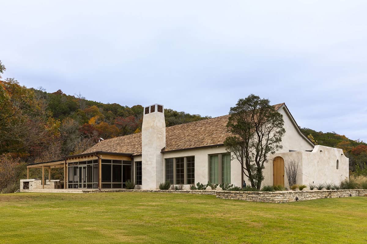 Full rear exterior of white stucco hacienda with cedar shake roof, screened porch, and wooded hill backdrop