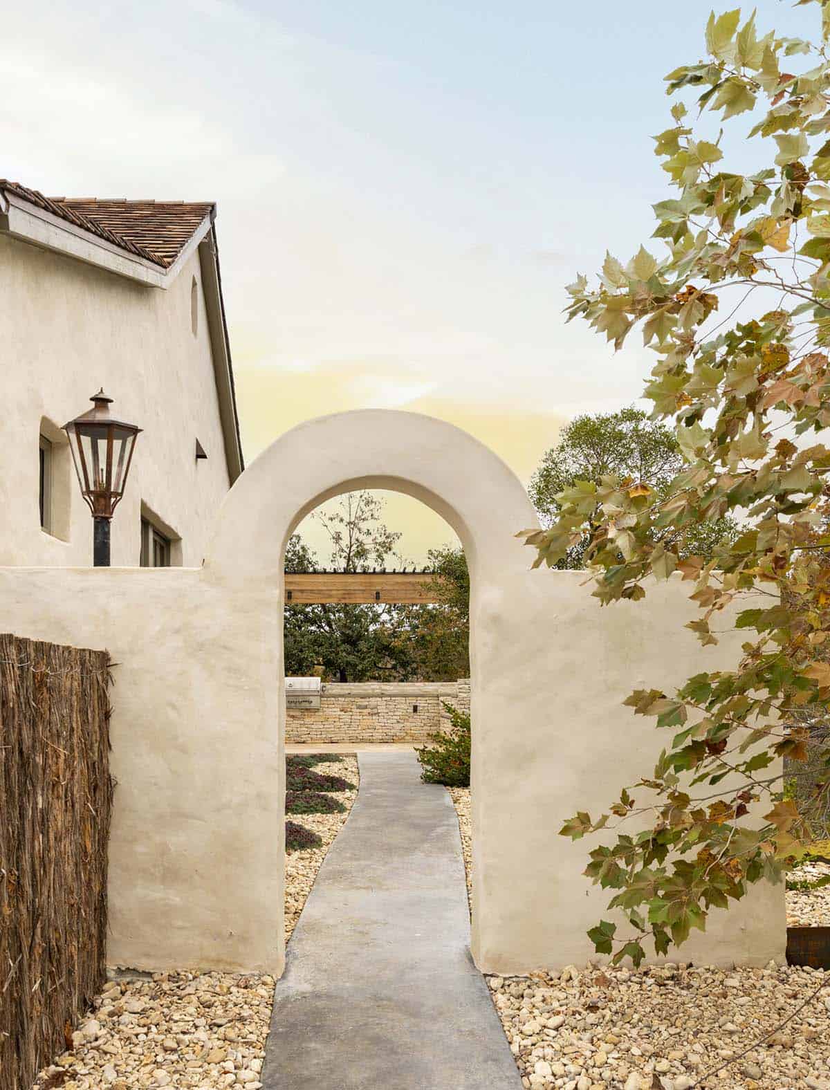 Plaster arched gateway leading to gravel courtyard path with copper gas lantern at sunset