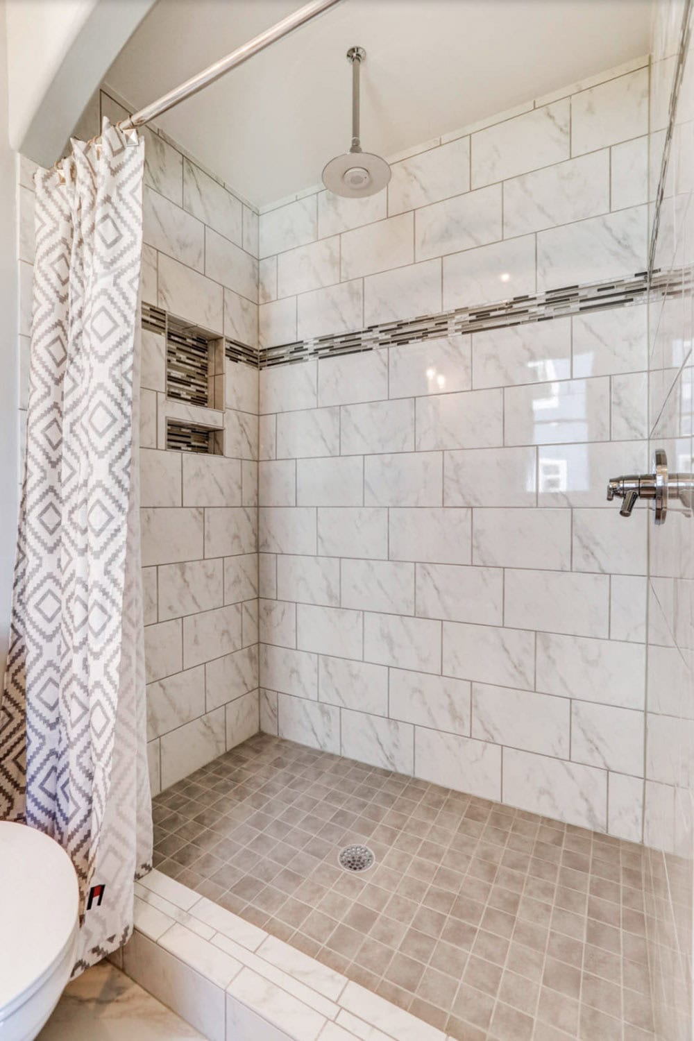 Walk-in shower with large marble-look subway tile, decorative mosaic border, built-in niches, and rain showerhead