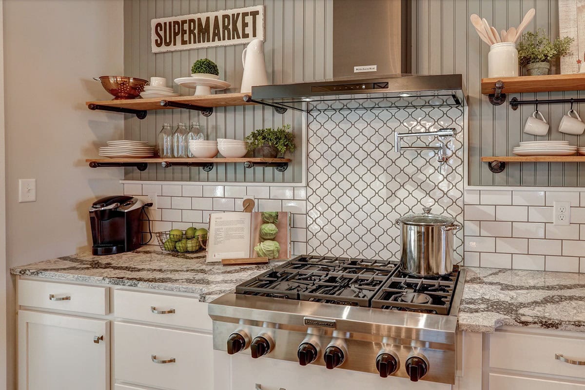 Farmhouse kitchen cooking wall with six-burner gas range, arabesque tile backsplash, open wood shelving, and range hood