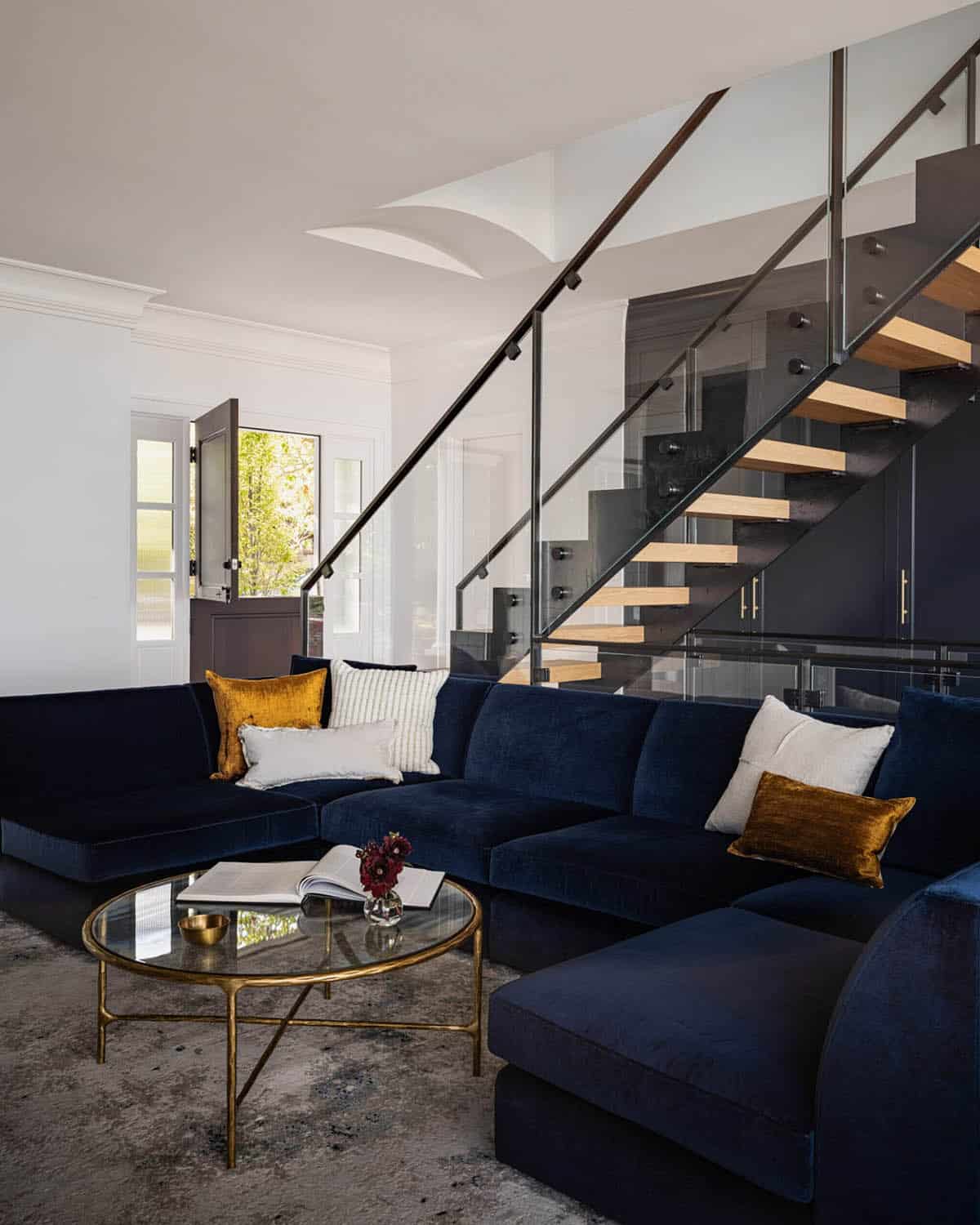 Living room with navy velvet sectional sofa, gold coffee table, and open floating staircase with glass railing