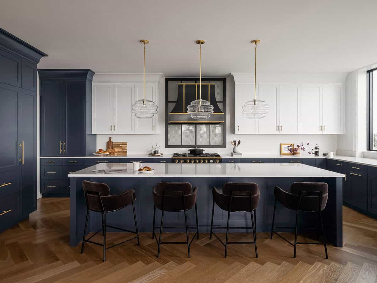 Navy and white two-tone kitchen with large island, bar stools, brass pendants, and herringbone hardwood floors