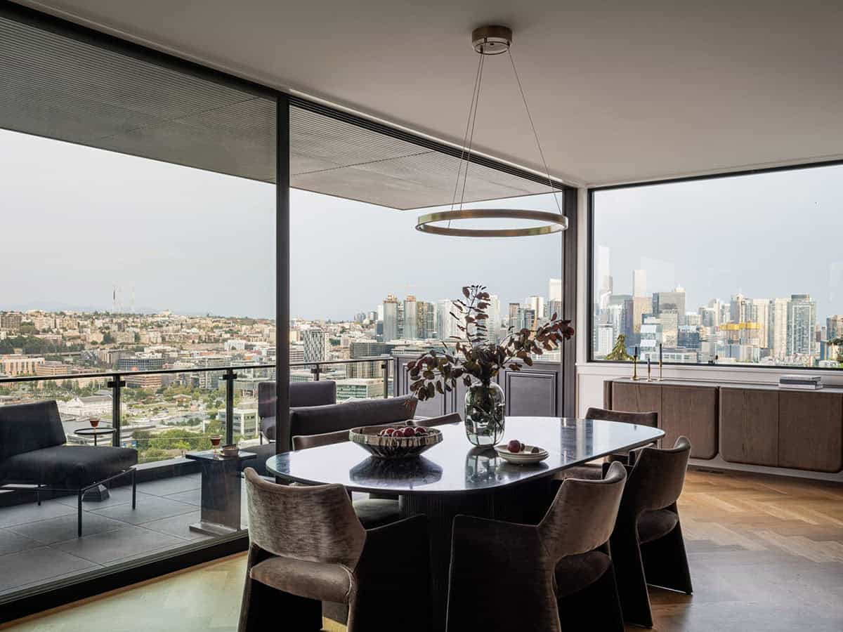 Dining area with dark oval table, velvet chairs, ring chandelier, and floor-to-ceiling city skyline views
