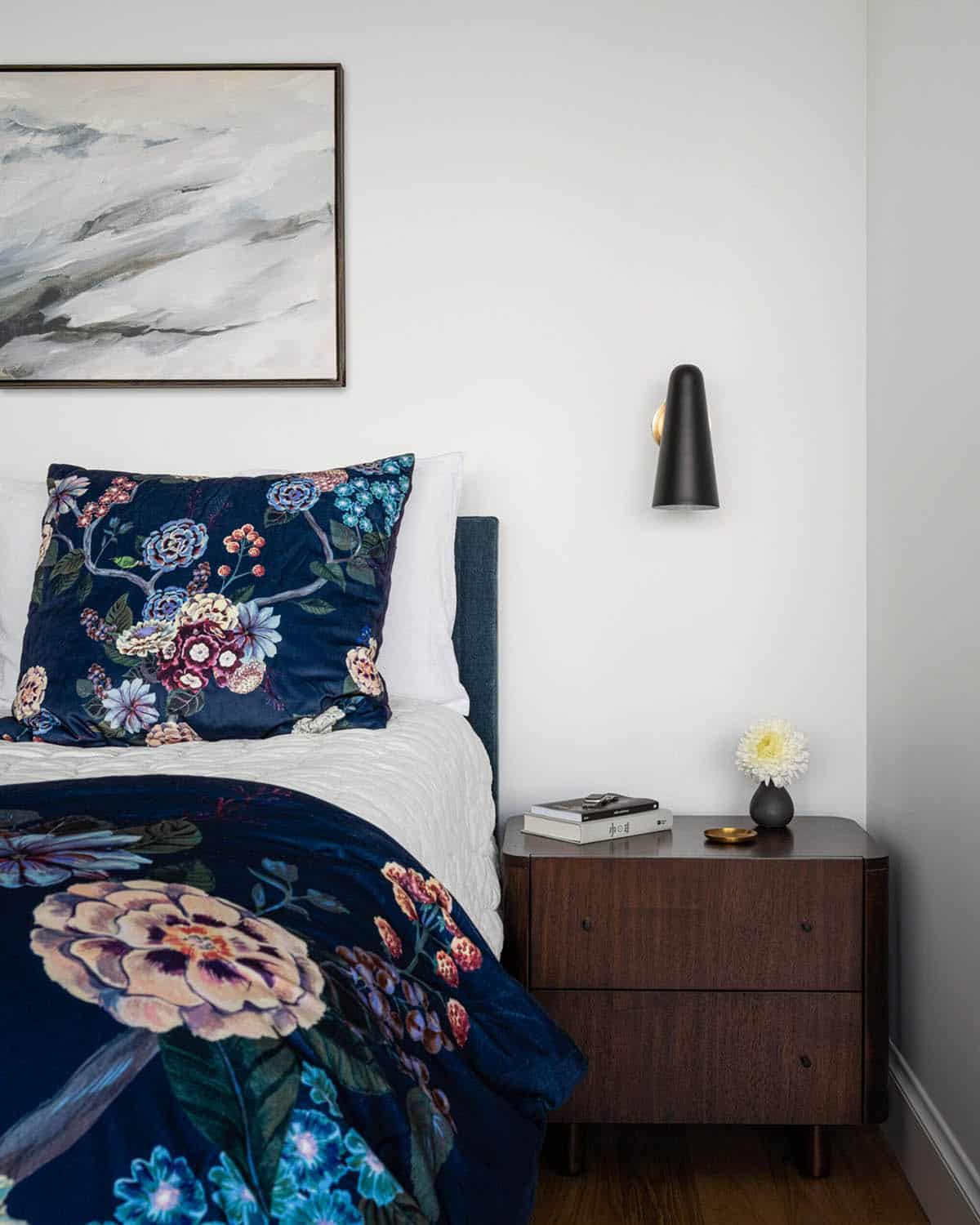 Bedroom detail with navy floral duvet, upholstered headboard, walnut nightstand, and wall sconce