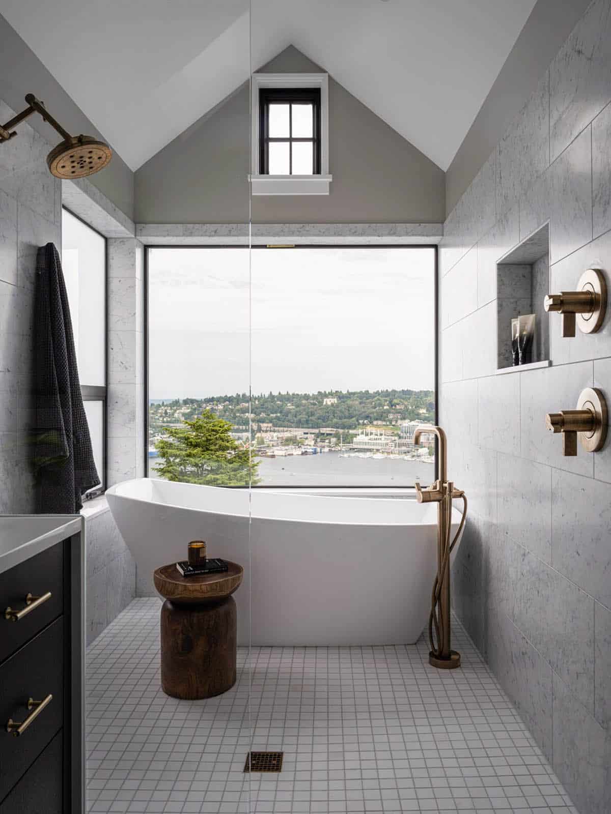 Freestanding soaking tub beside floor-to-ceiling window with lake and city views in marble-tiled bathroom