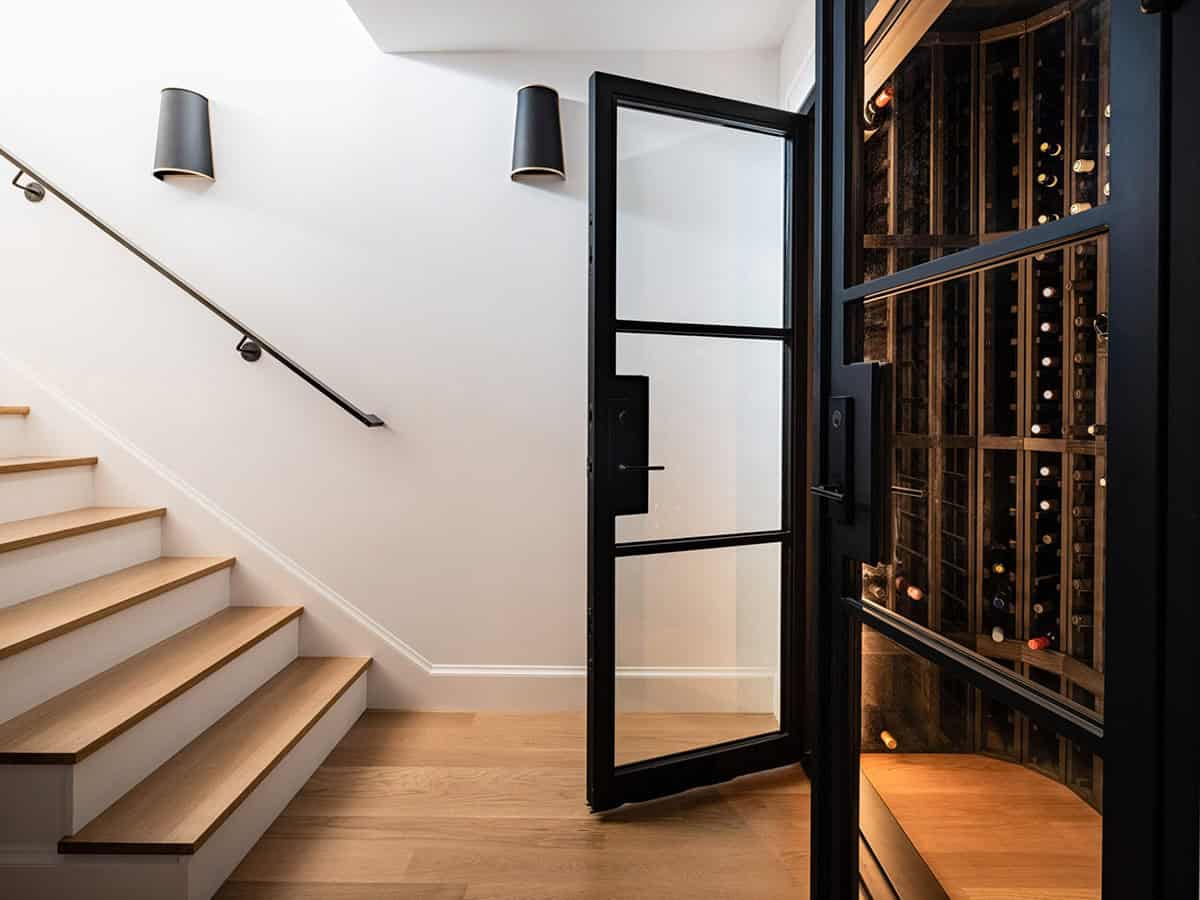 Glass-door wine cellar beside staircase with black steel frame and wall-mounted sconces
