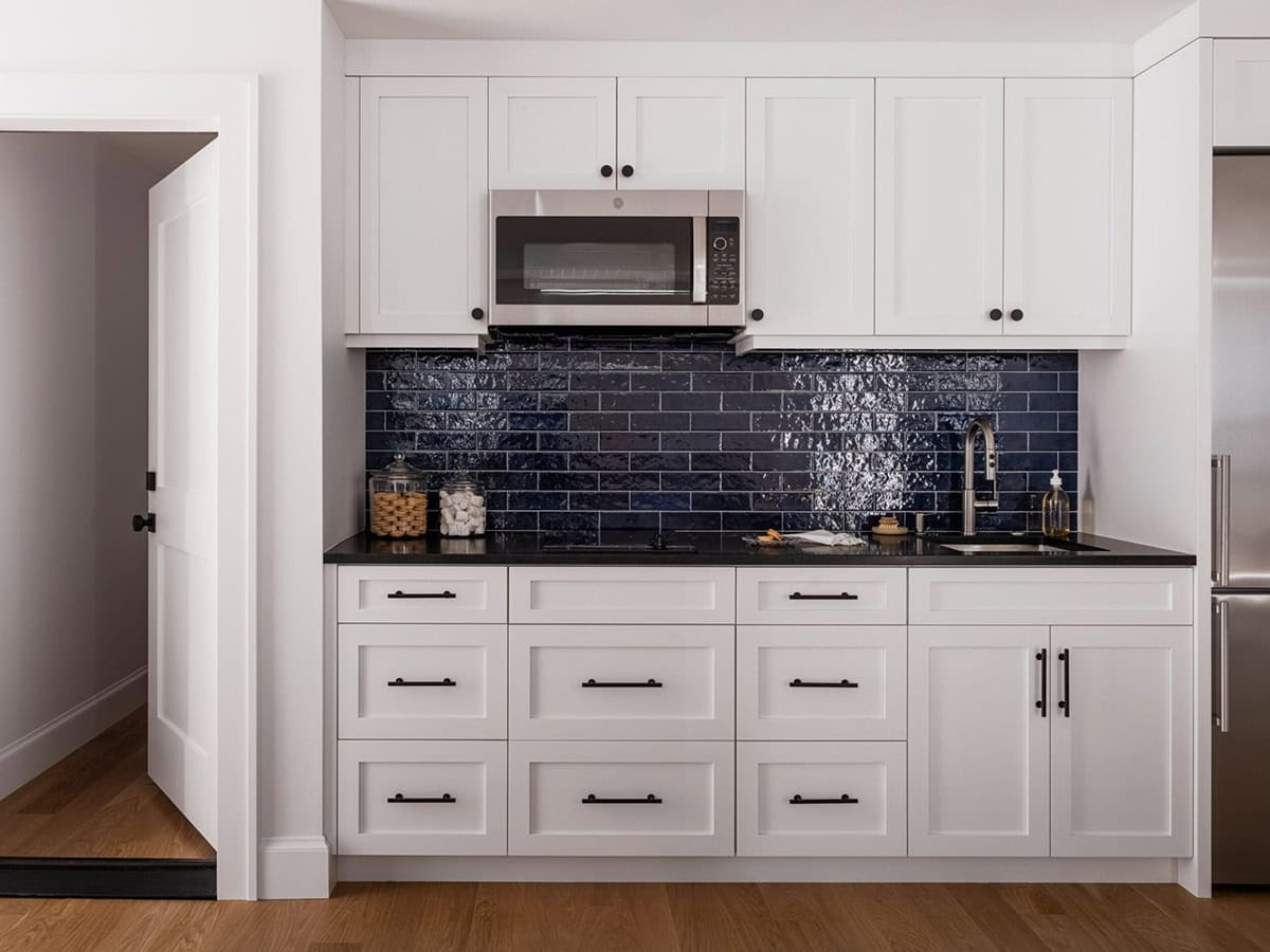 White shaker kitchen cabinetry with navy glazed tile backsplash, black hardware, and built-in microwave