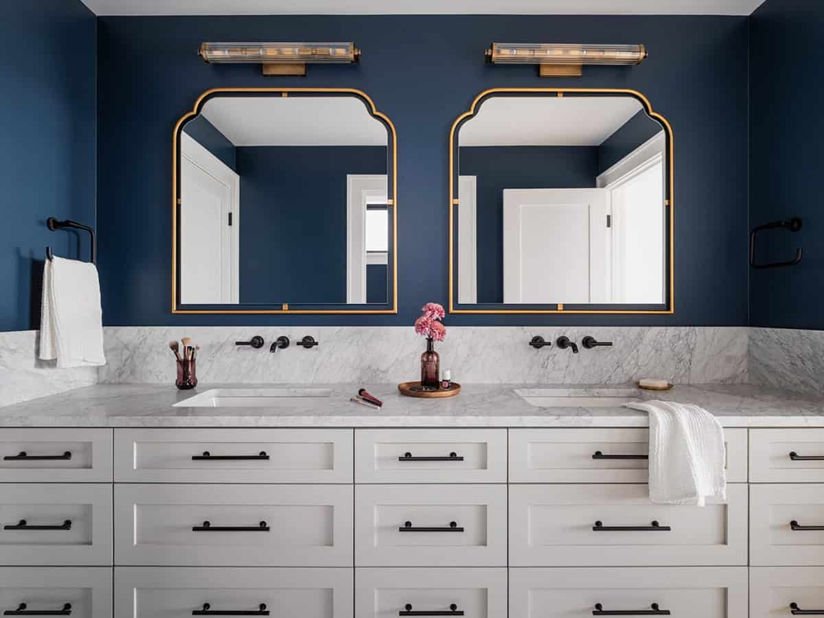 Double vanity bathroom with navy blue walls, dual gold-framed arch mirrors, and marble countertop