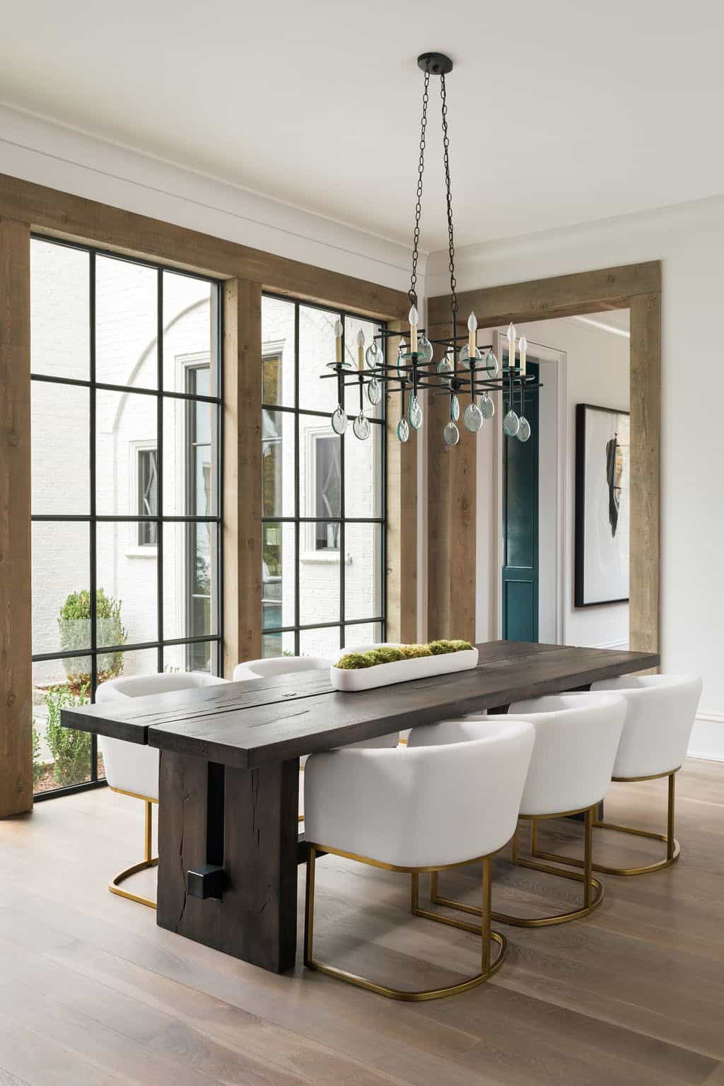 Modern English country breakfast room with dark wood trestle table, white velvet chairs with brass legs and cedar-framed steel windows in Georgia