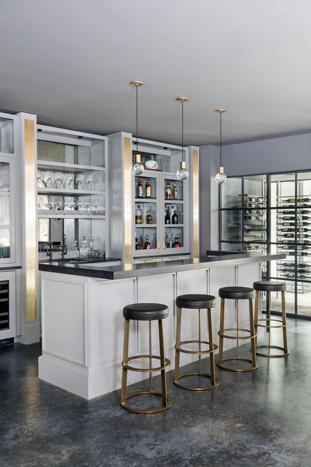 Modern English country basement bar with white cabinetry, dark granite countertop, brass bar stools and glass wine cellar in Georgia