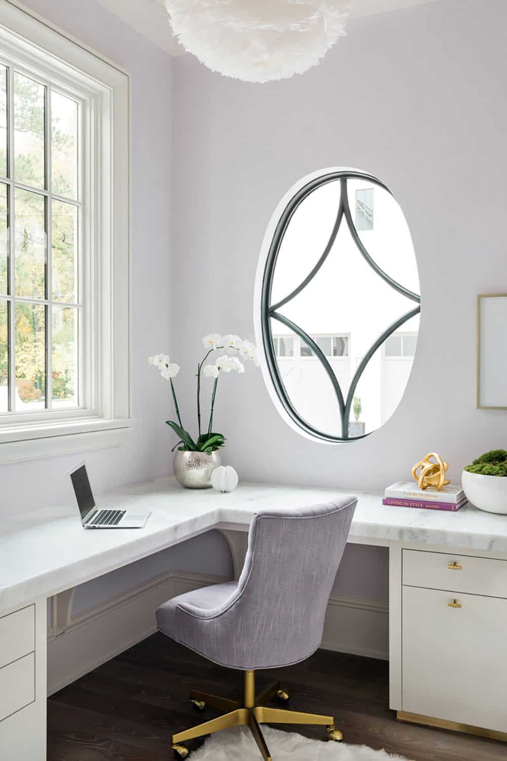 Modern English country house home office with marble L-shaped desk, oval steel-framed window, lavender walls and brass desk chair in Georgia