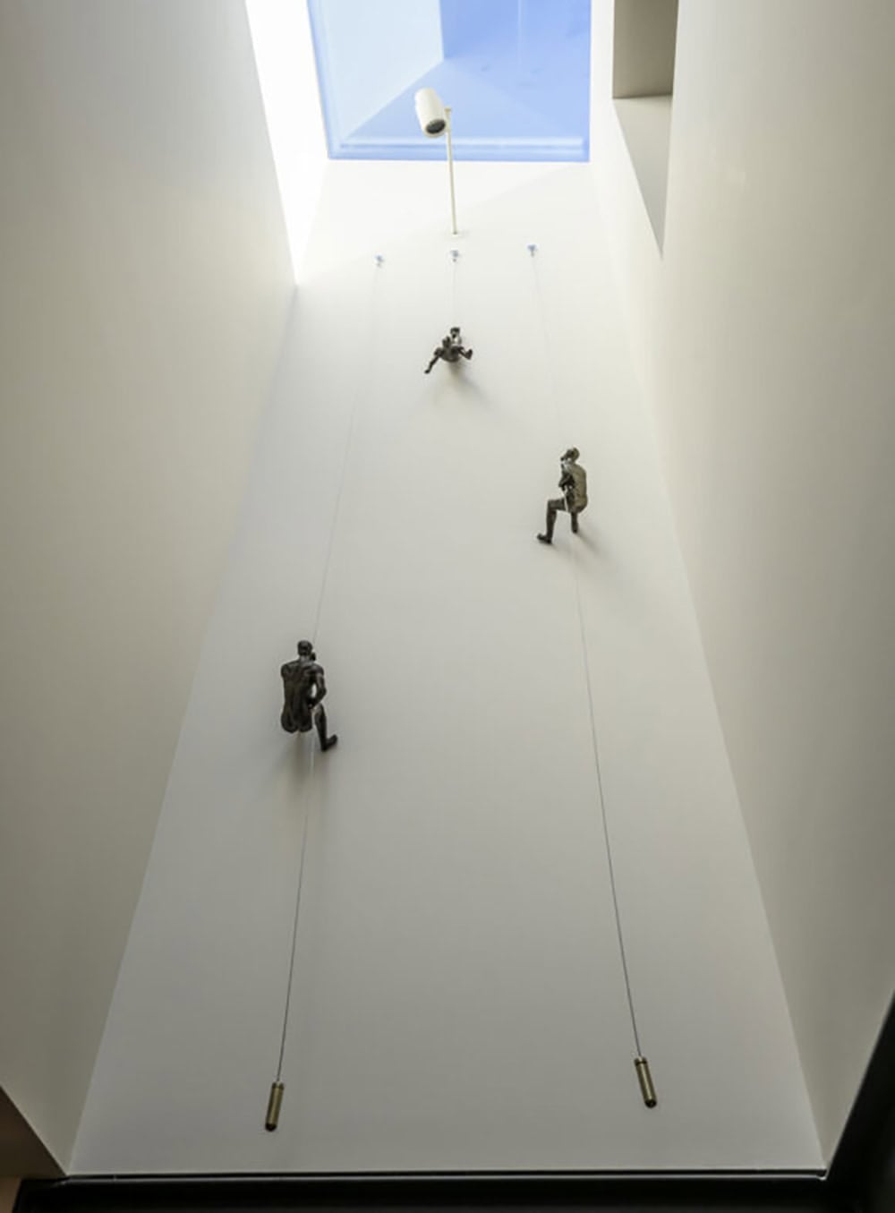 Three bronze climbing figure sculptures ascending stairwell wall beneath skylight